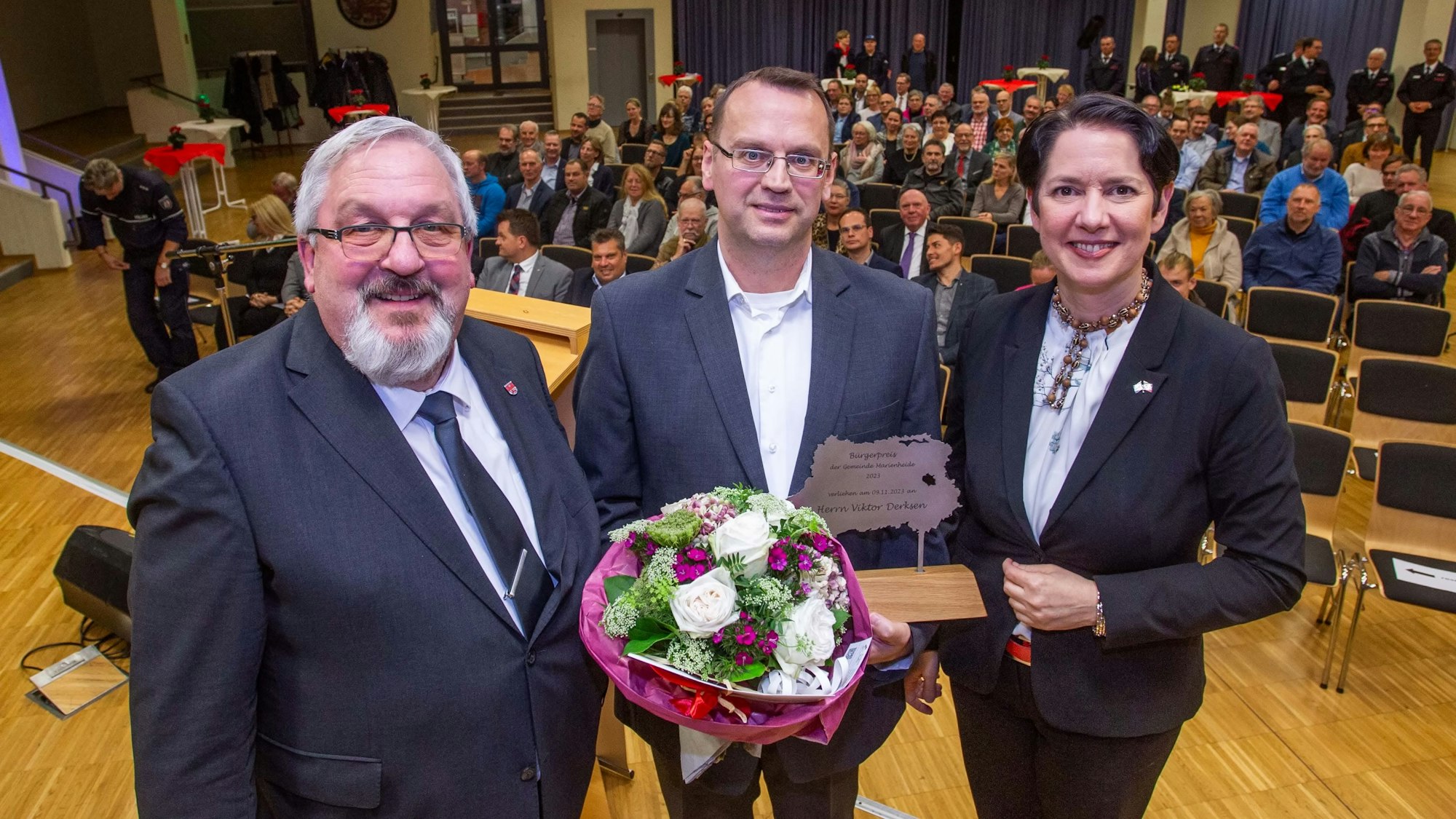 Zwei Männer und eine Frau stehen zusammen. Der Mann in der Mitte hält einen Bürgerpreis und Blumen in den Händen. Im Hintergrund ist Publikum zu sehen.