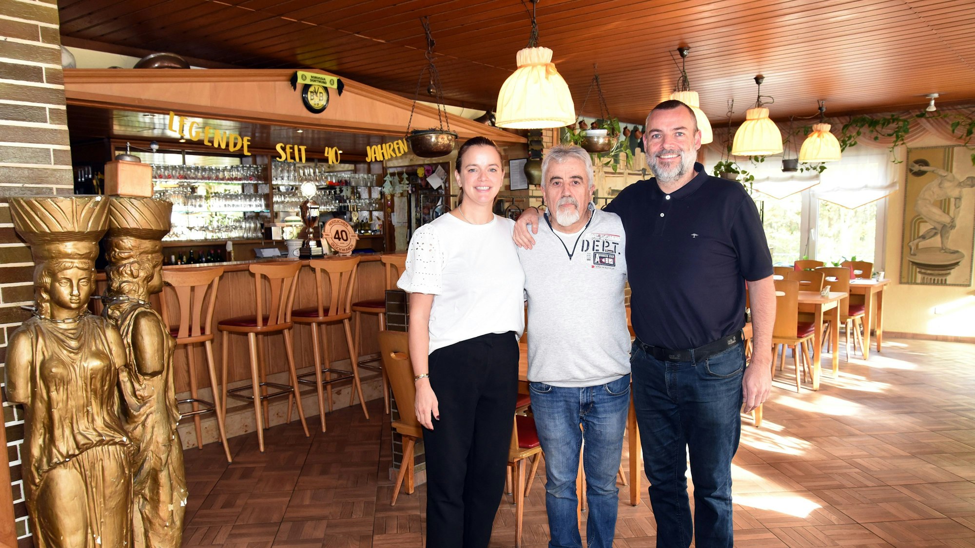 Zwei Männer und eine Frau stehen Arm in Arm im Gastraum eines Restaurants.