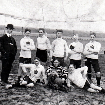 Eine alte Schwarz-Weiß-Fotografie zeigt Fußballer in Trikots und Männer mit Anzug und Melonen-Hüten.