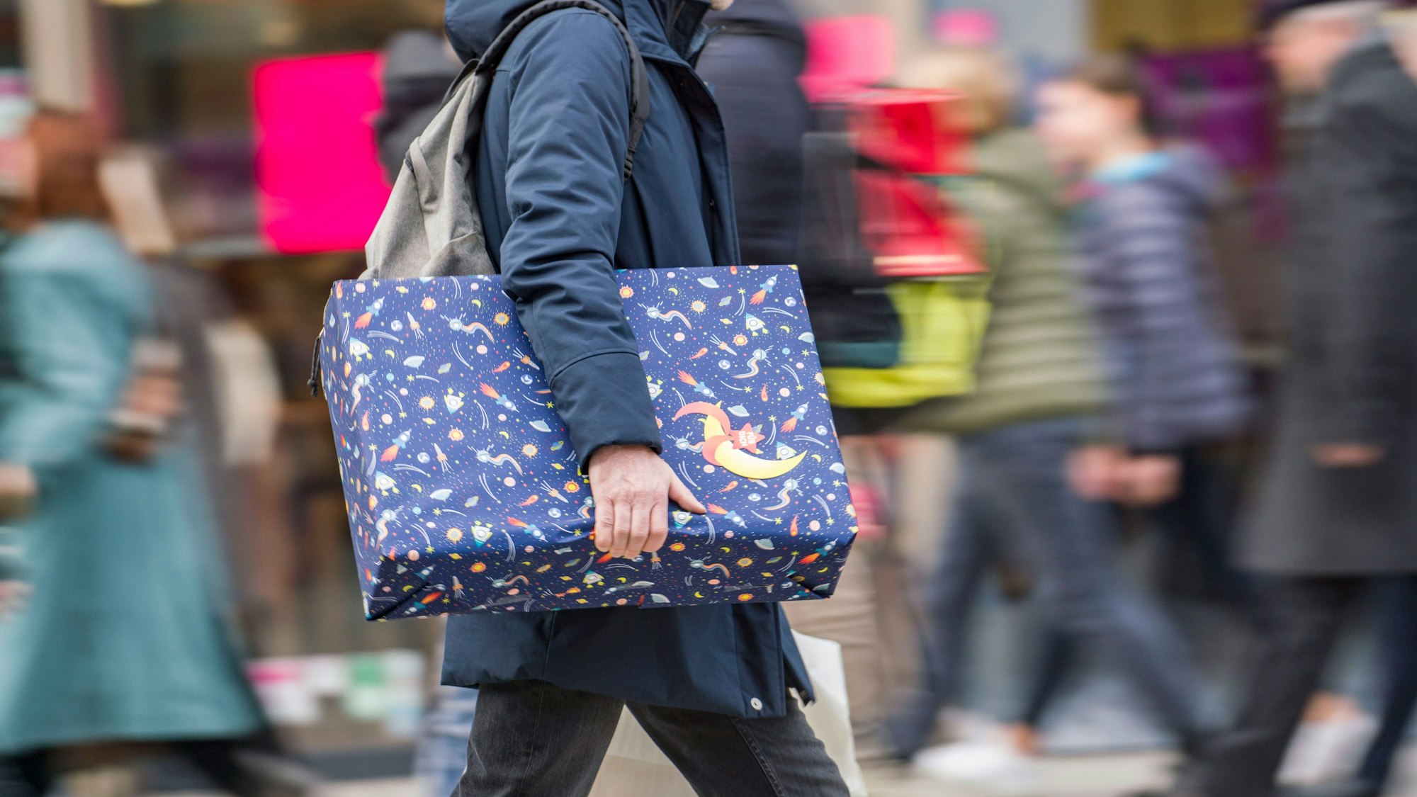 Ein Mann eilt mit einem verpackten Geschenk zwischen den anderen Passanten die Einkaufsmeile entlang.