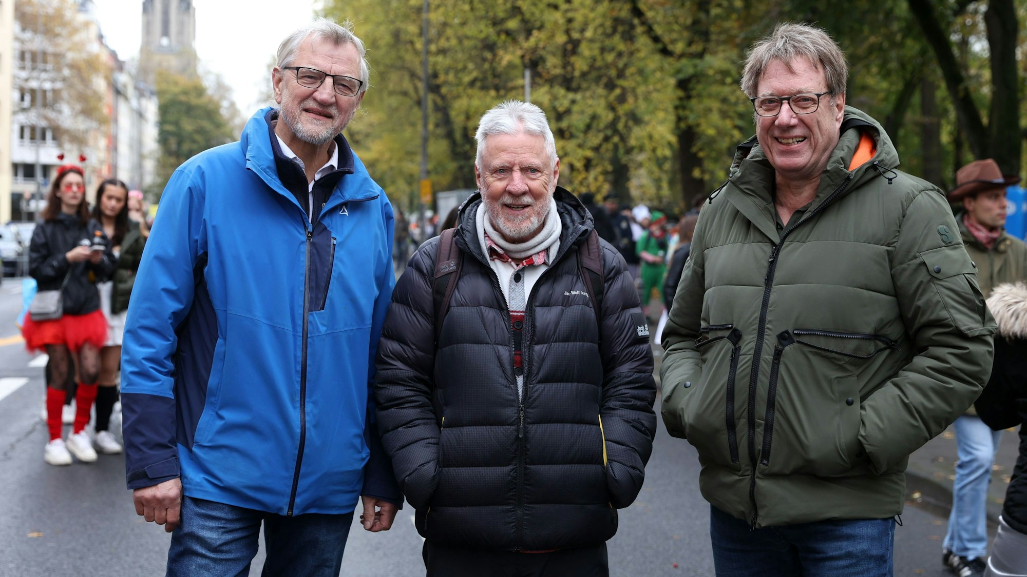 Bezirksbürgermeister Andreas Hupke, Klaus Adrian von der Bürgervereinigung Rathenauplatz und Michael Neumann stehen am 11.11. auf der Straßen im Kwartier Latäng, im Hintergrund sind verkleidete Menschen zu sehen.