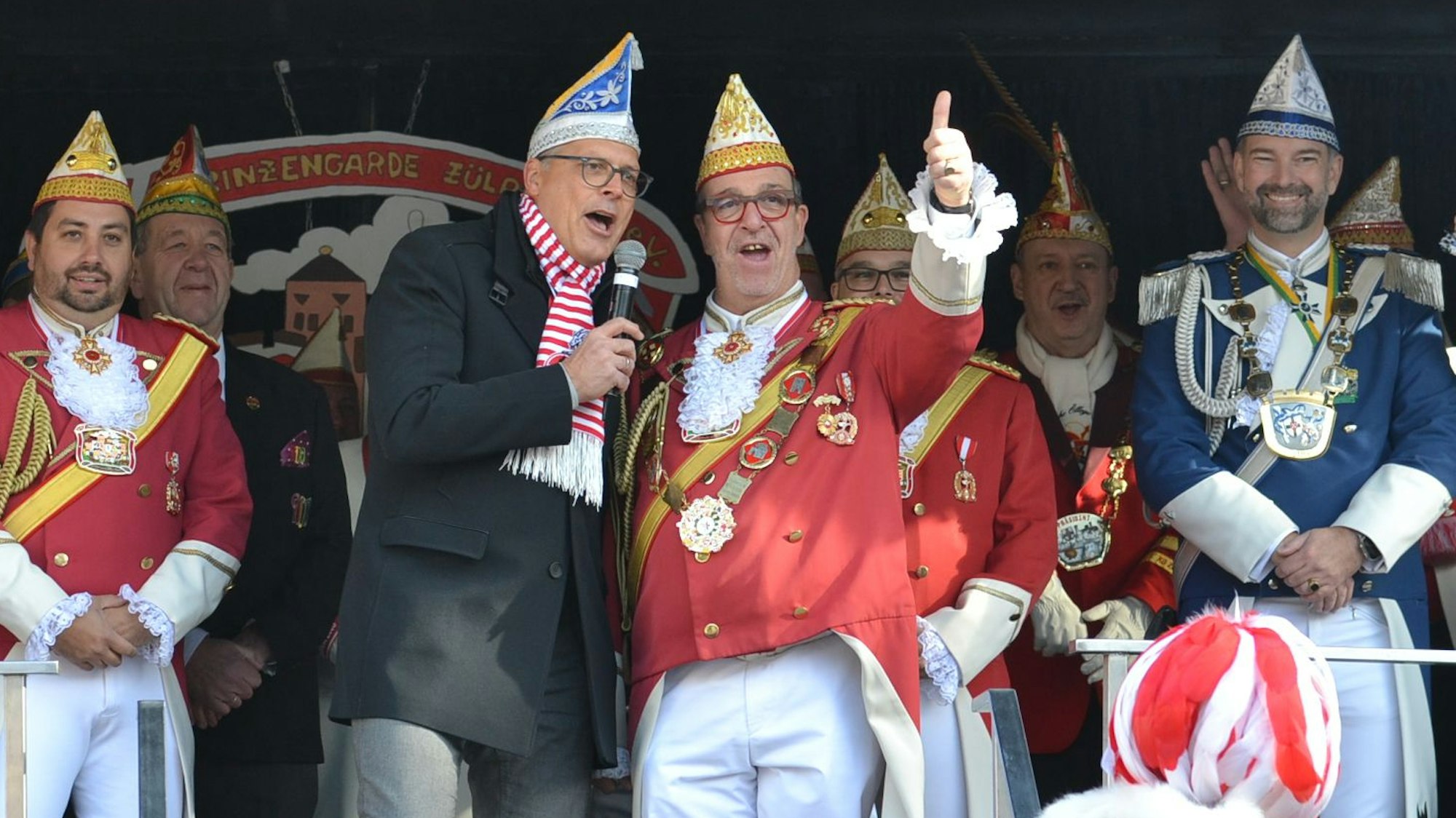 Zülpichs Bürgermeister Ulf Hürtgen trägt Narrenkappe und steht mit Mitgliedern von Prinzengarde und Blauen Funken auf der Bühne.