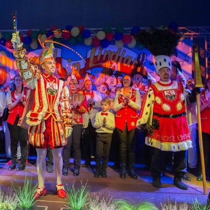 Ein Karnevals-Dreigestirn steht auf einer bunt geschmückten Bühne in einem Festsaal.