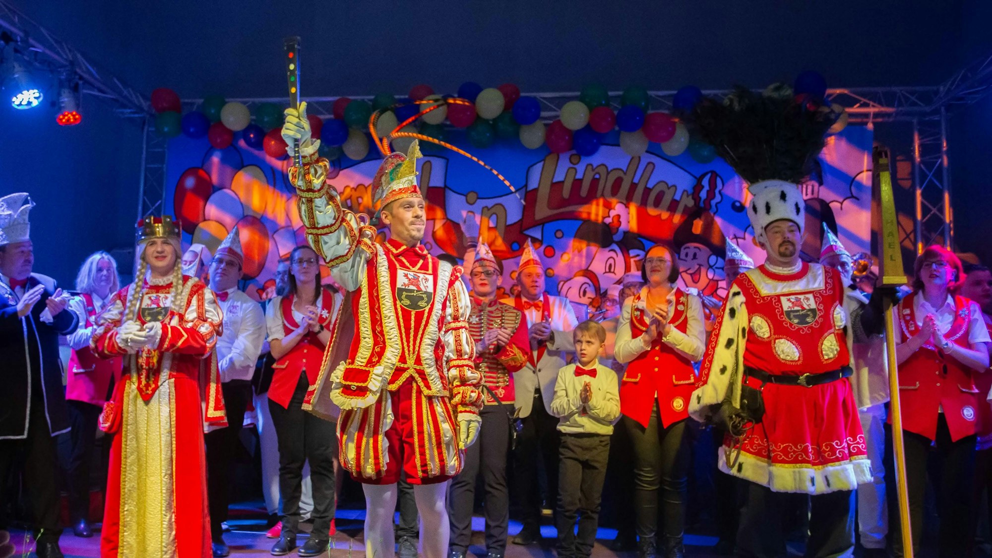 Ein Karnevals-Dreigestirn steht auf einer bunt geschmückten Bühne in einem Festsaal.