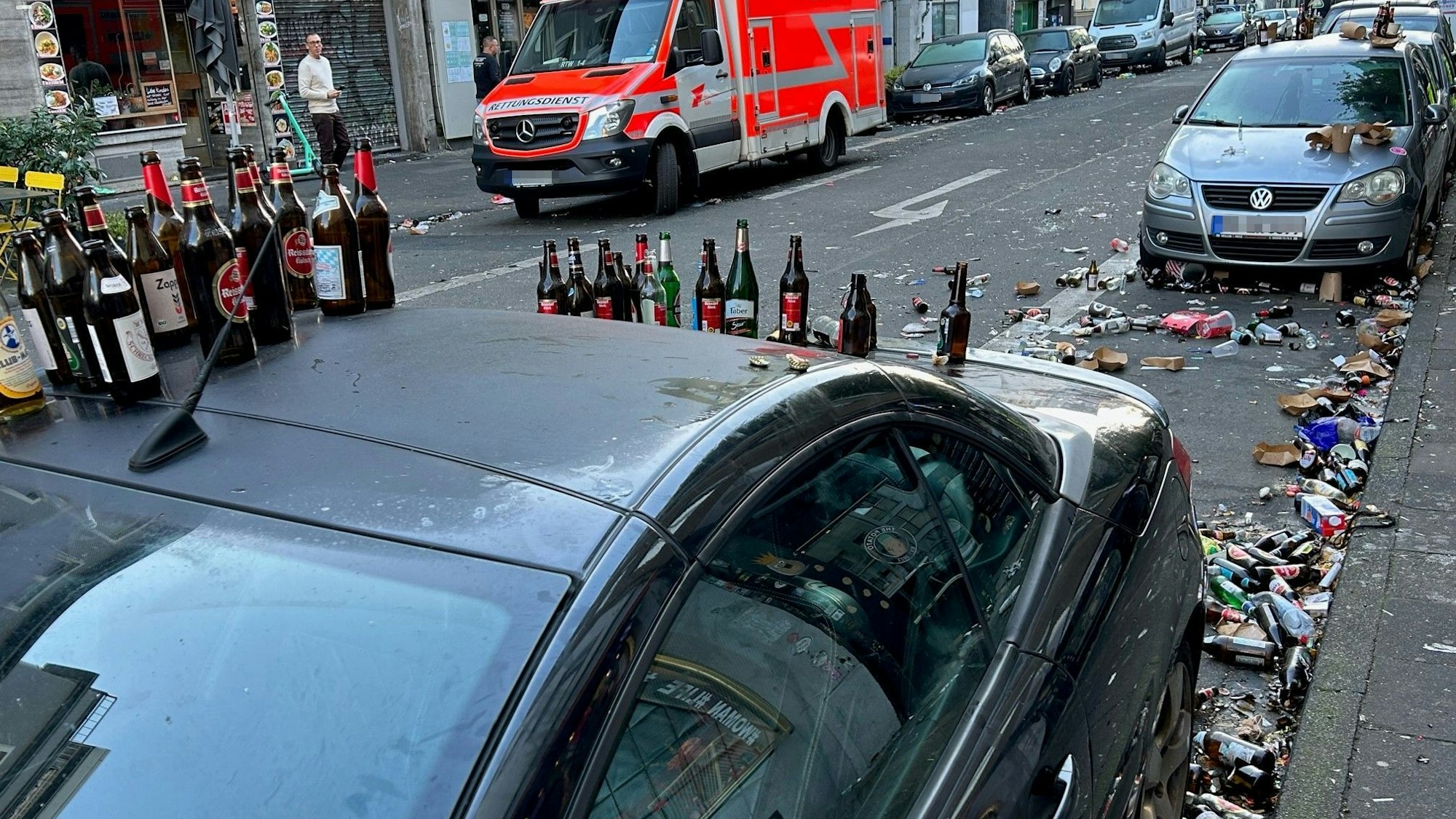 Bierflaschen stehen auf dem Dach eines geparkten Autos, auch auf der Straße liegen Berge von Glasflaschen.