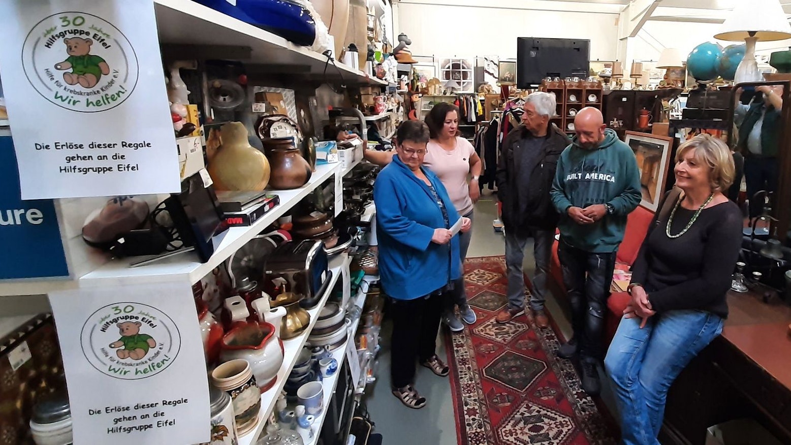 Hilfsgruppenvorsitzender Willi Greuel (Mitte) besuchte den Dauerflohmarkt von Maria Hamacher (2.v.l.) in Kall. Weitere Besucher betrachten die mit vielen Artikeln vollgestellten Regale.