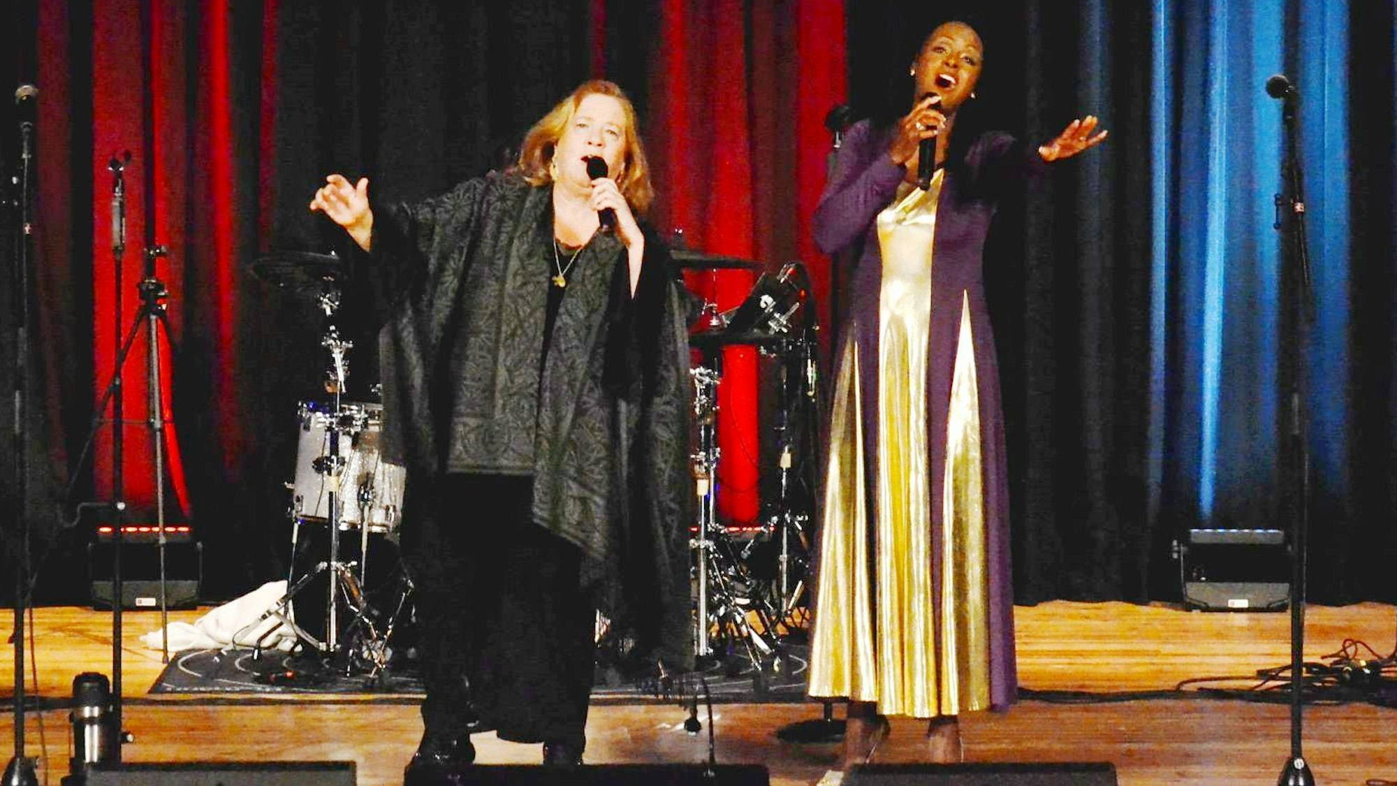 Im Vordergrund auf der Bühne die schwarz gekleidete Kathy Kelly, begleitet von einer weiteren Sängerin im violett- und goldfarbenem Kleid.