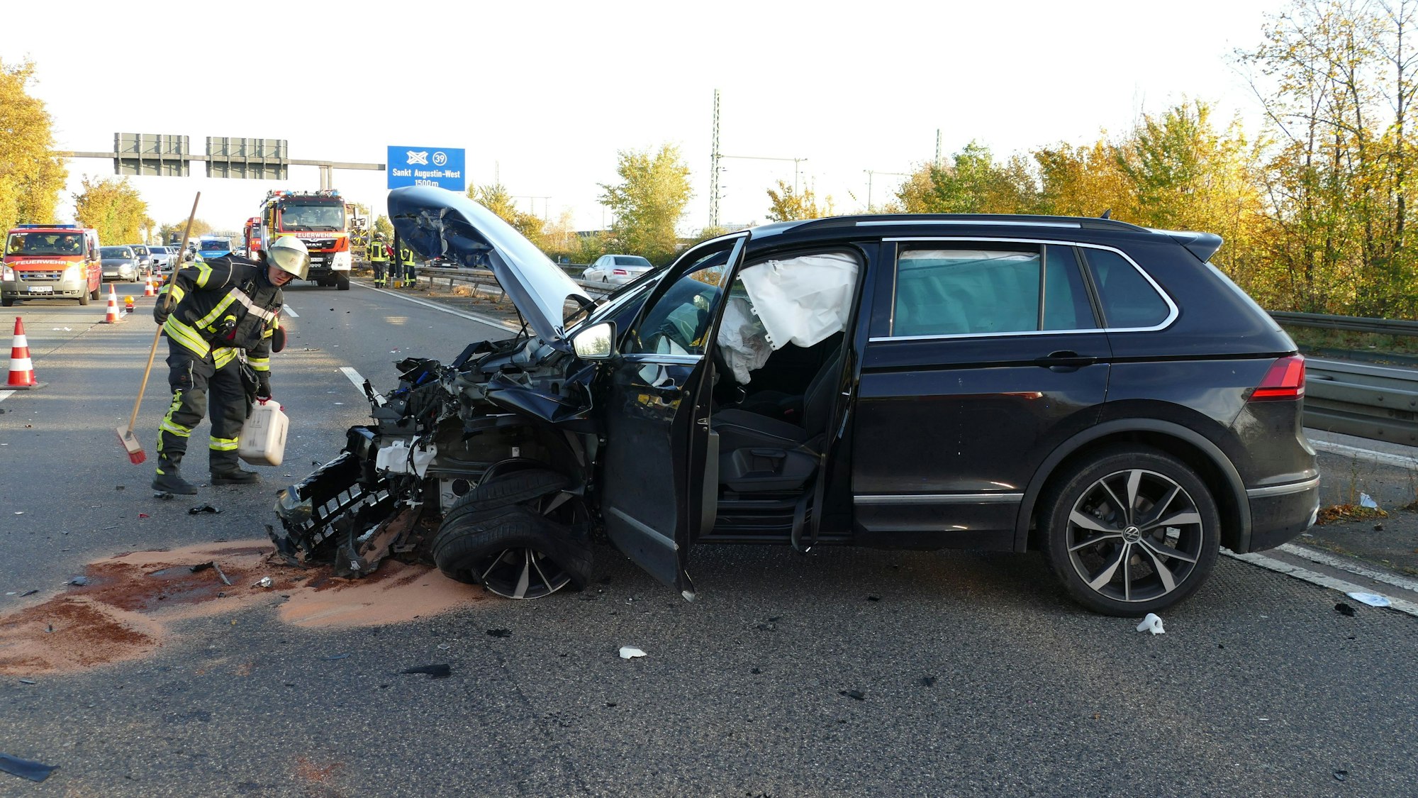 Ein zerstörter VW Tiguan auf der Autobahn. Im Hintergrund Fahrzeuge der Feuerwehr und des Rettungsdienstes. Ein Feuerwehrmann verstreut Bindemittel auf der Fahrbahn.