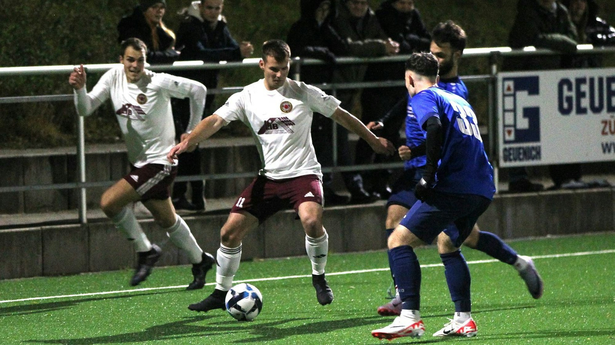 Lommersums Marcel Ebel hat den Ball und wird von zwei Gegenspielern bedrängt. Ein Mitspieler hinterläuft Ebel.