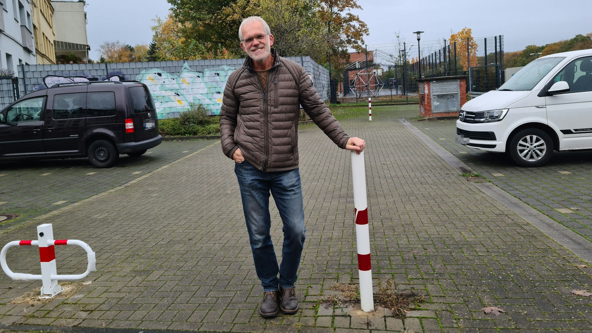 Ein Mann steht auf einem Parkplatz auf einen rot-weißen Poller gestützt. Auf dem Parkplatz sind viele Plätze frei.