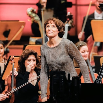 Helene Grimaud steht von ihrem Flügel auf und lächelt wegen des Applauses des Publikums. Im Hintergrund weitere Mitglieder des Orchesters.