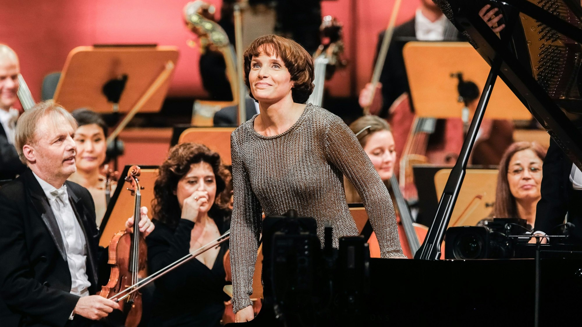 Helene Grimaud steht von ihrem Flügel auf und lächelt wegen des Applauses des Publikums. Im Hintergrund weitere Mitglieder des Orchesters.