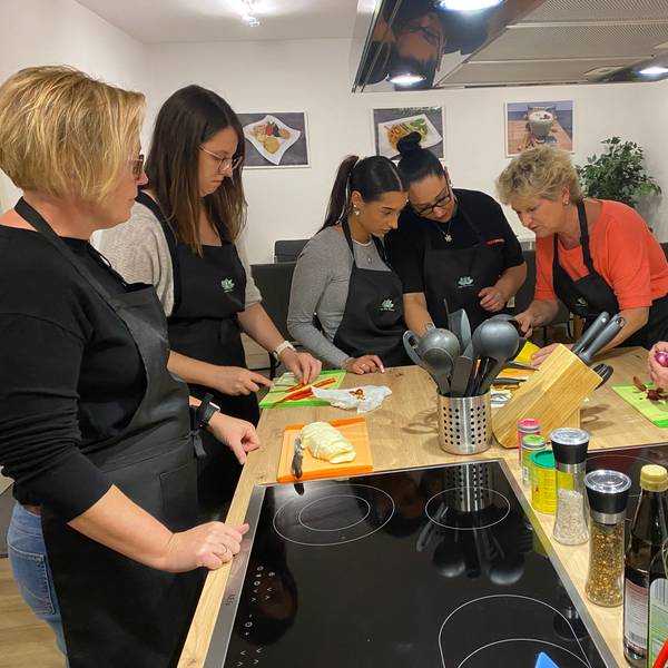 Sechs Frauen mit Schürzen stehen um eine Kochinsel und schnibbeln frische Zutaten wie Paprika und Zwiebeln.