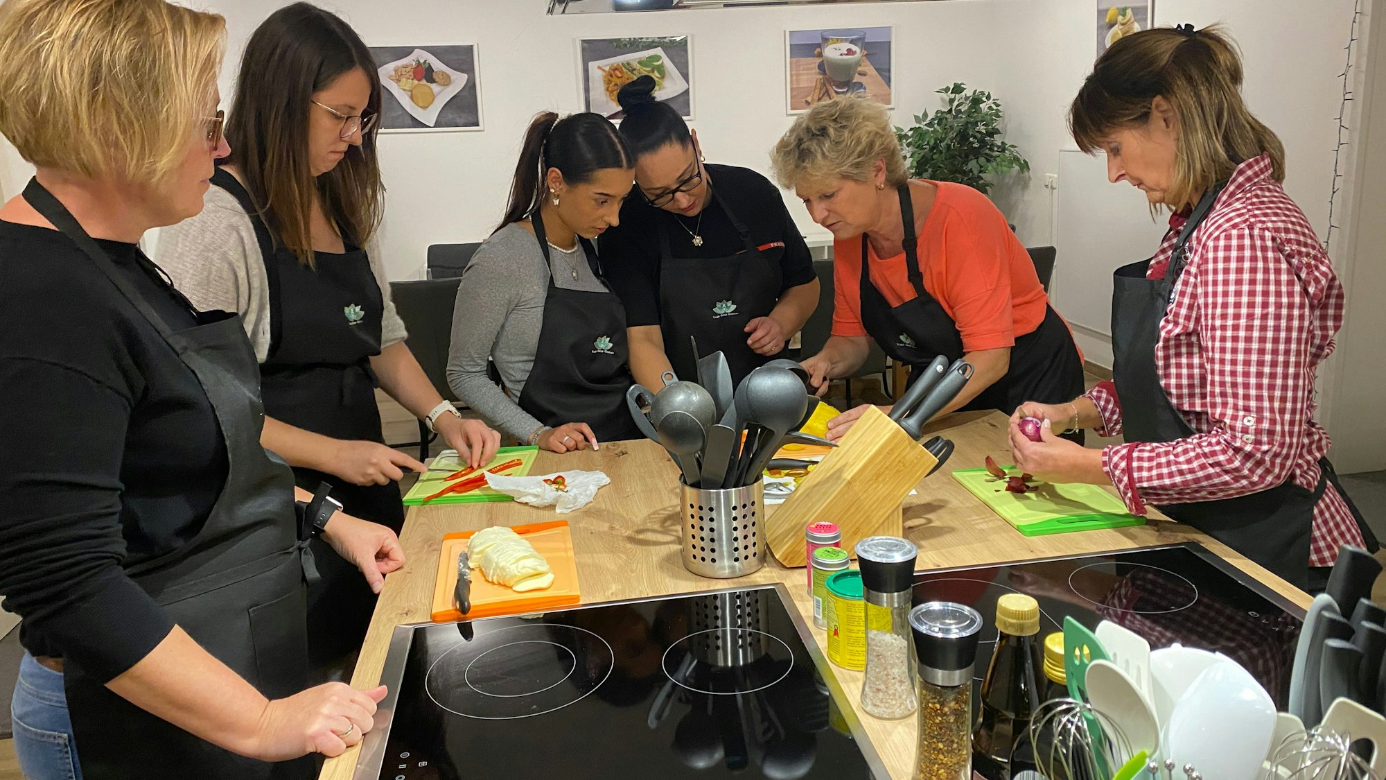 Sechs Frauen mit Schürzen stehen um eine Kochinsel und schnibbeln frische Zutaten wie Paprika und Zwiebeln.