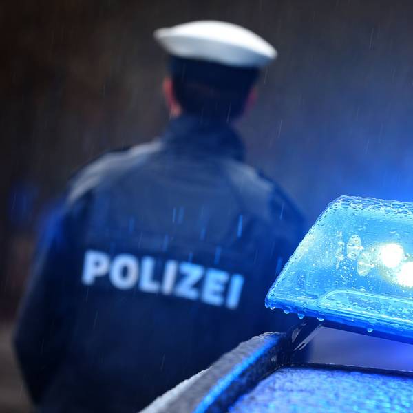 Ein Polizist steht vor einem Streifenwagen dessen Blaulicht aktiviert ist.