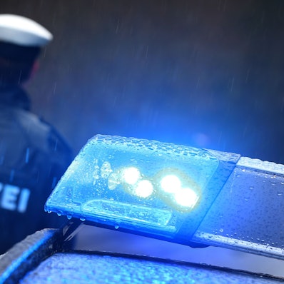Ein Polizist steht vor einem Streifenwagen dessen Blaulicht aktiviert ist.