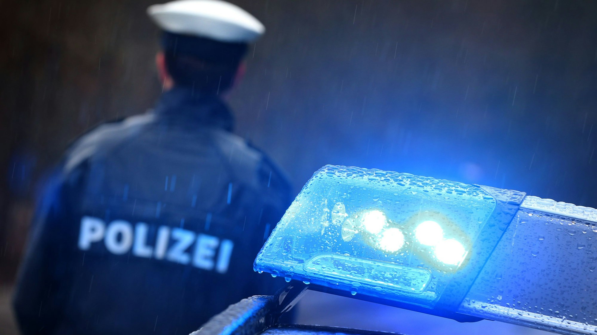 Ein Polizist steht im Regen vor einem Streifenwagen, dessen Blaulicht aktiviert ist.