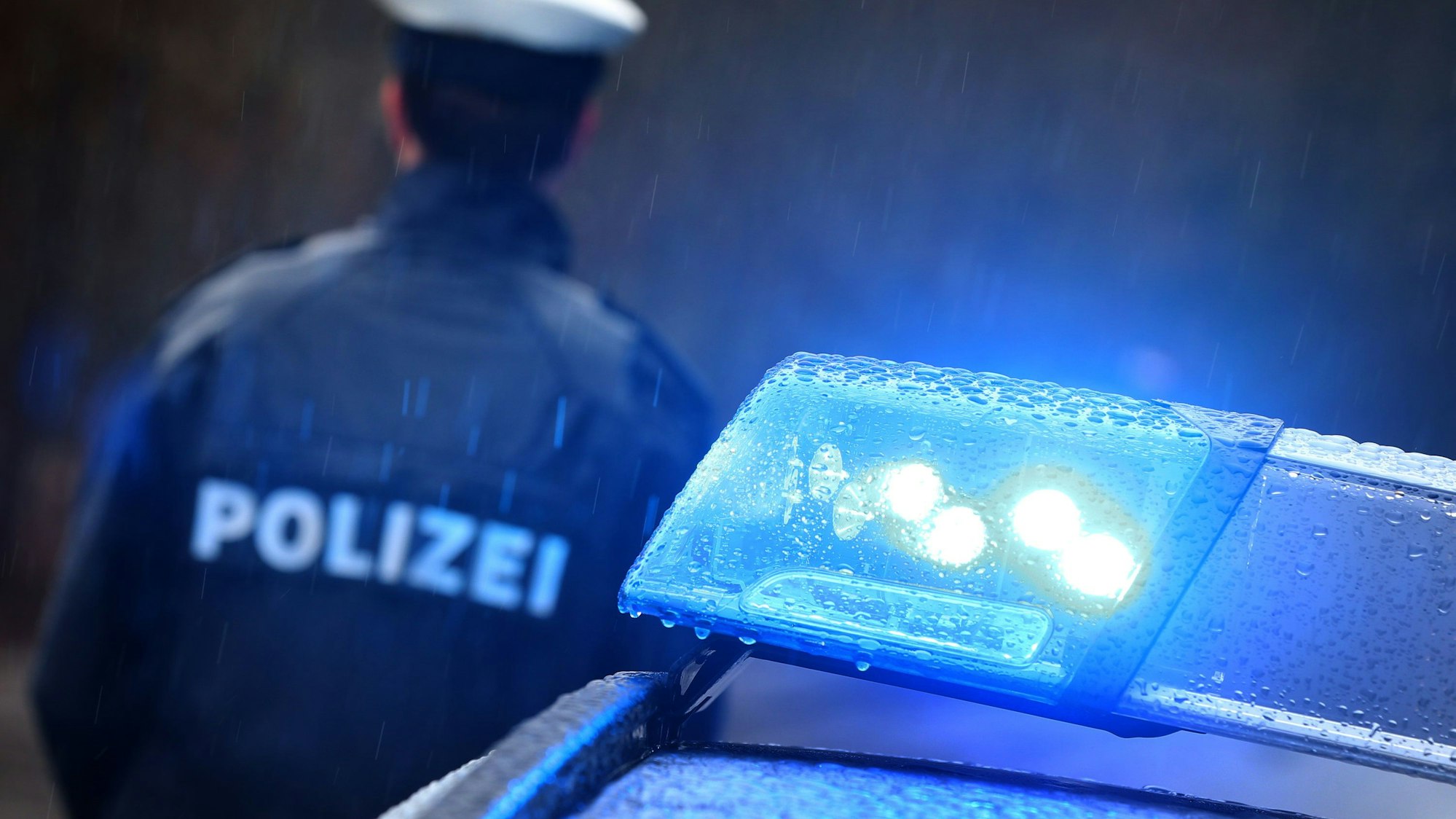 Ein Polizist steht im Regen vor einem Streifenwagen dessen Blaulicht aktiviert ist.