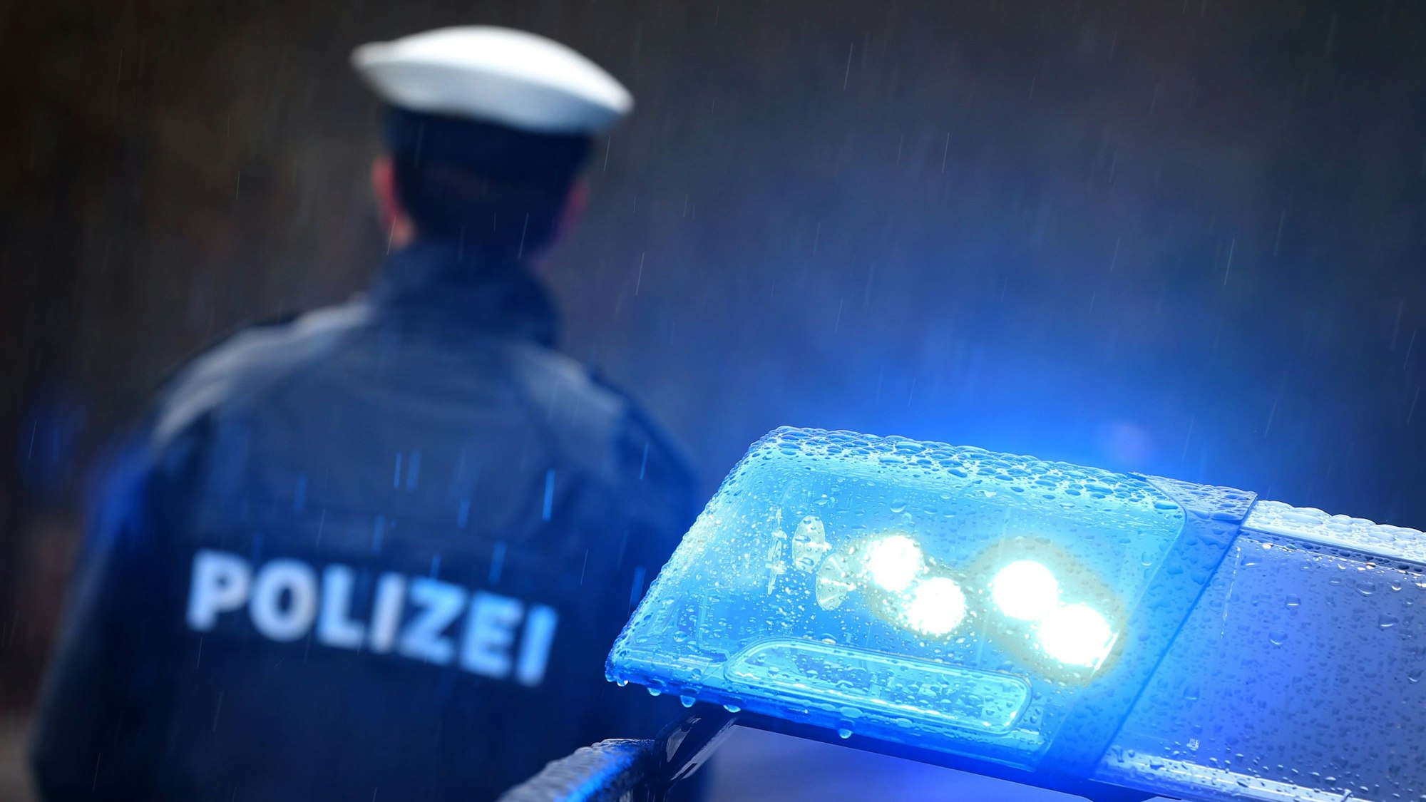 Ein Polizist steht vor einem Streifenwagen dessen Blaulicht aktiviert ist.