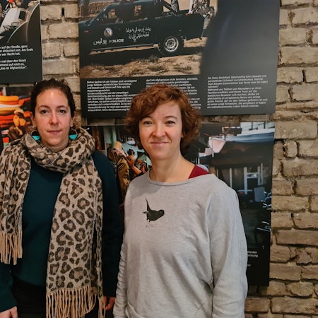 Zwei junge Frauen stehen vor Bildern einer Ausstellung, auf einem ist ein Mädchen zu sehen, das einen schweren Sack trägt.