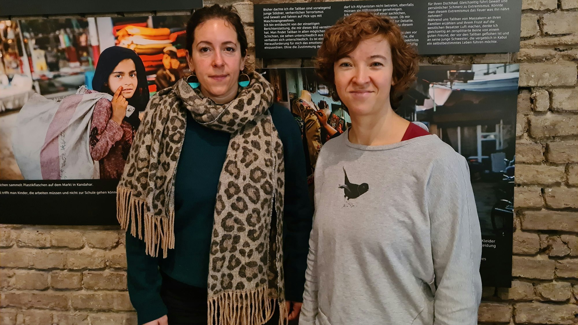 Zwei Frauen stehen nebeneinander vor einer Wand, an der Fotografien von Frauen hängen.