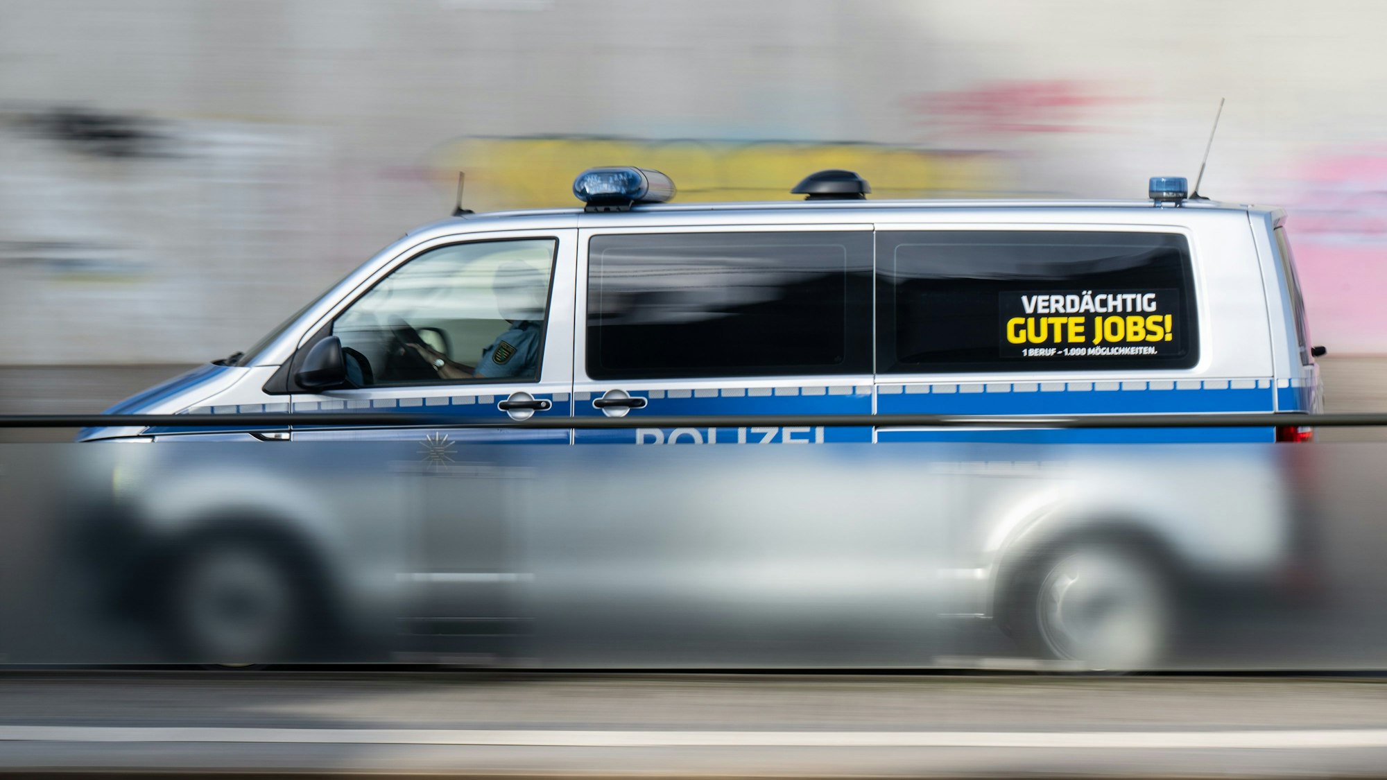 Ein Polizeiauto mit der Aufschrift ·Verdächtig gute Jobs!· fährt eine Straße entlang. (Aufnahme mit langer Belichtungszeit)