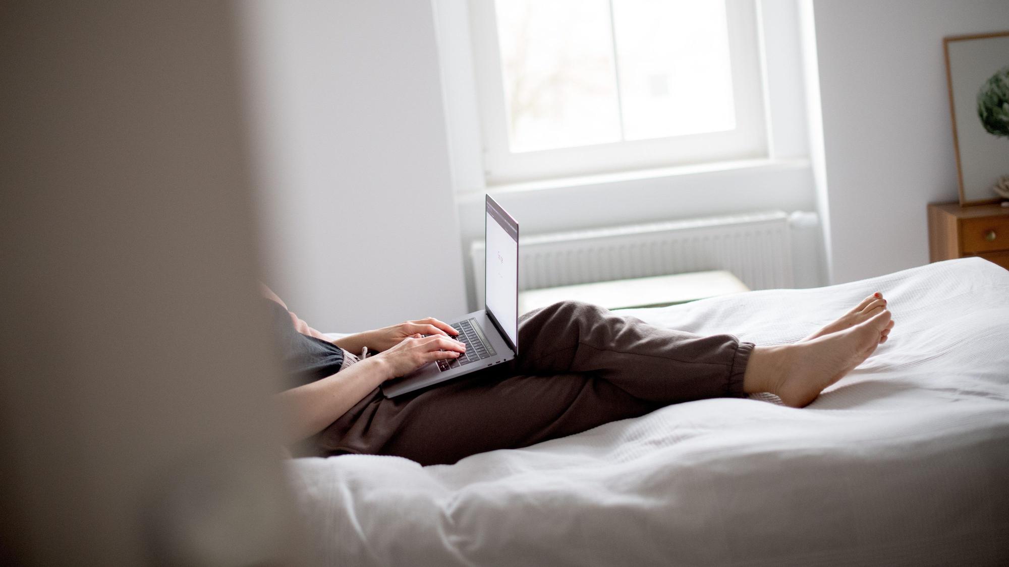 Ein Mensch liegt in Jogginghose auf dem Bett und arbeitet am Laptop