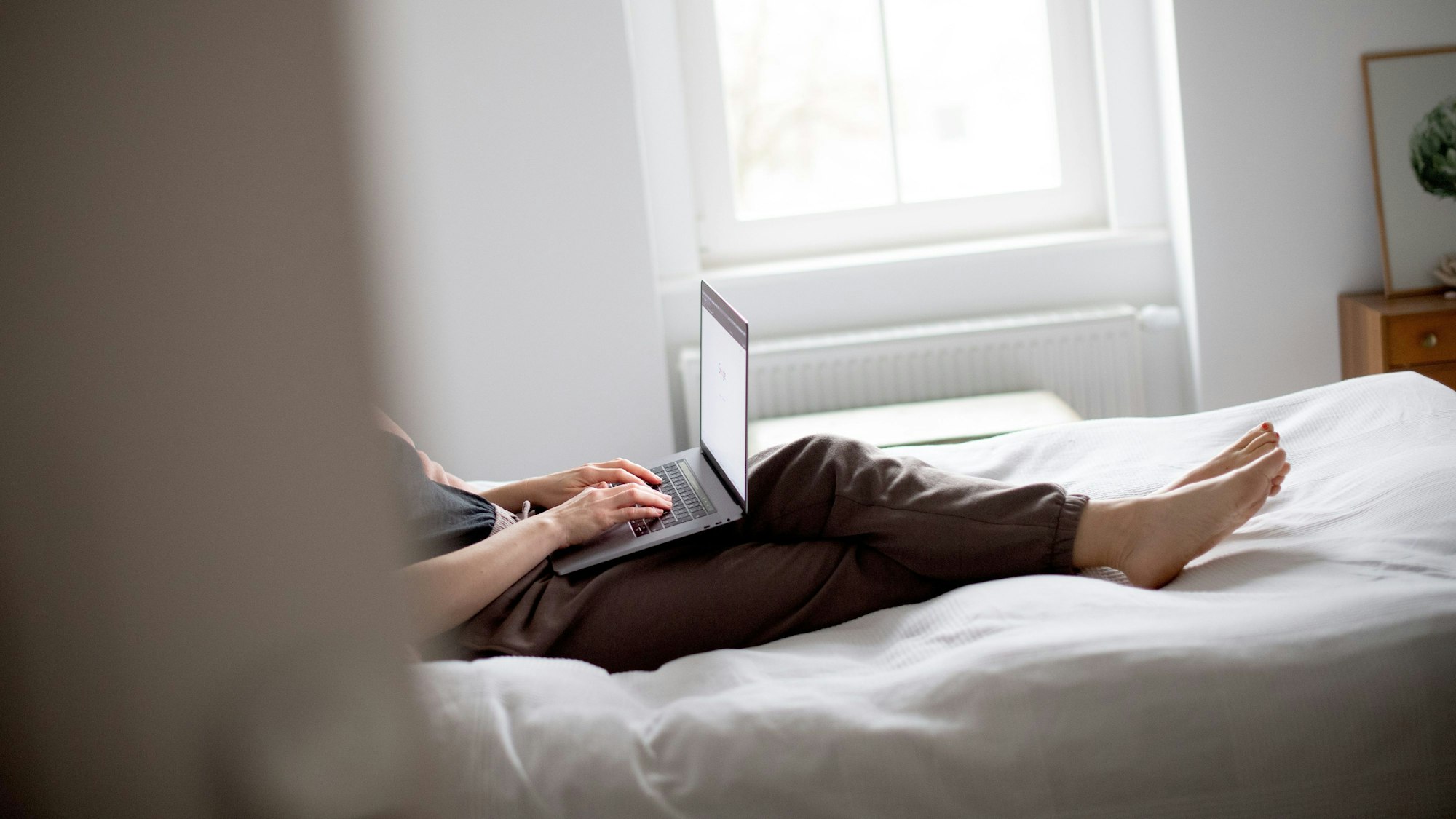 Ein Mensch liegt in Jogginghose auf dem Bett und arbeitet am Laptop