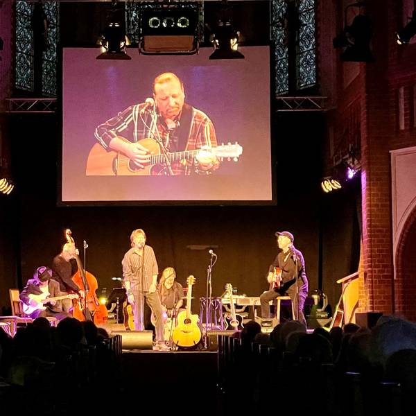 Gerd Köster (Mitte), Helmut Krumminga (rechts) und weitere Weggefährten stehen bei der Gedenkfeier von Frank Hocker in der Kulturkirche in Nippes gemeinsam auf der Bühne. Hinter ihnen ist das Bild von dem verstorbenen Frank Hocker auf einer großen Leinwand zu sehen.