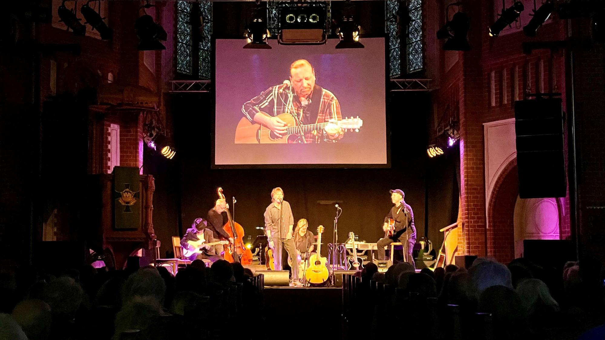 Gerd Köster (Mitte), Helmut Krumminga (rechts) und weitere Weggefährten stehen bei der Gedenkfeier von Frank Hocker in der Kulturkirche in Nippes gemeinsam auf der Bühne. Hinter ihnen ist das Bild von dem verstorbenen Frank Hocker auf einer großen Leinwand zu sehen.