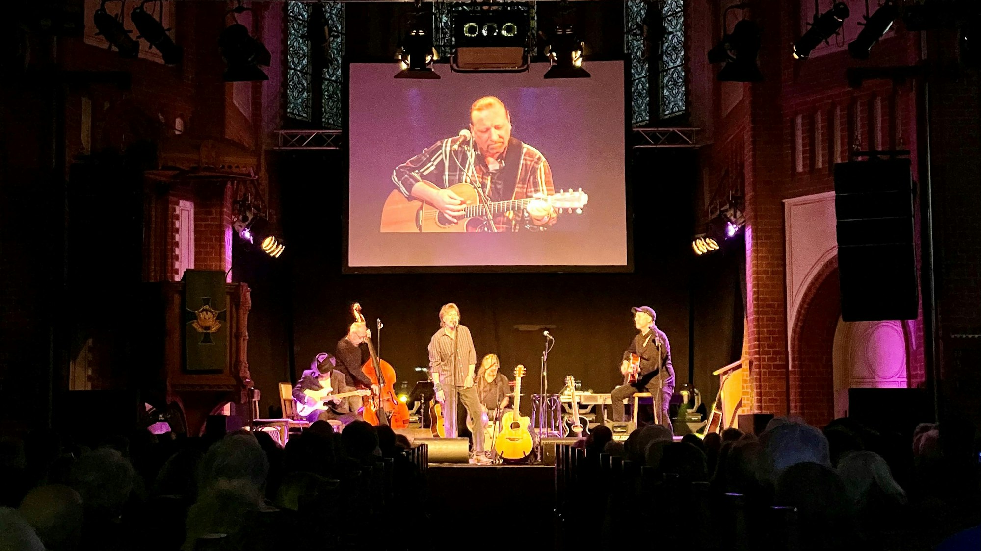 Gerd Köster (Mitte), Helmut Krumminga (rechts) und weitere Weggefährten stehen bei der Gedenkfeier von Frank Hocker in der Kulturkirche in Nippes gemeinsam auf der Bühne. Hinter ihnen ist das Bild von dem verstorbenen Frank Hocker auf einer großen Leinwand zu sehen.