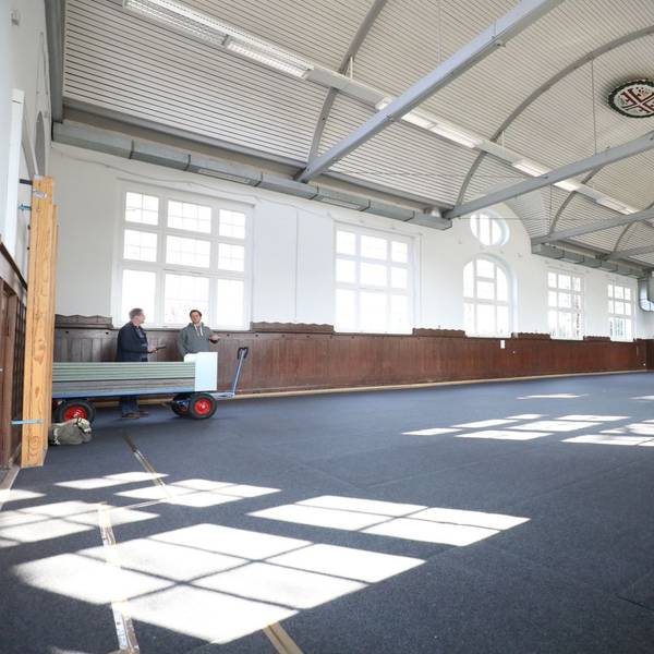 In einer fast leeren Turnhalle mit blauem Boden stehen am linken Rand zwei Personen.