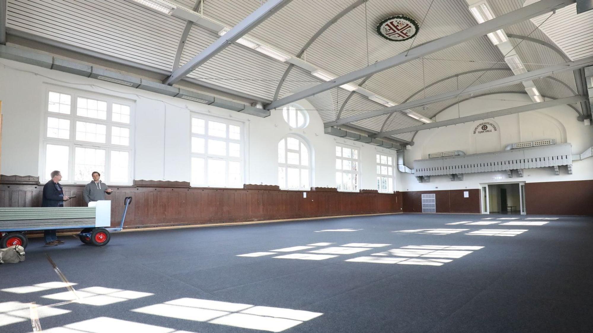 In einer fast leeren Turnhalle mit blauem Boden stehen am linken Rand zwei Personen.