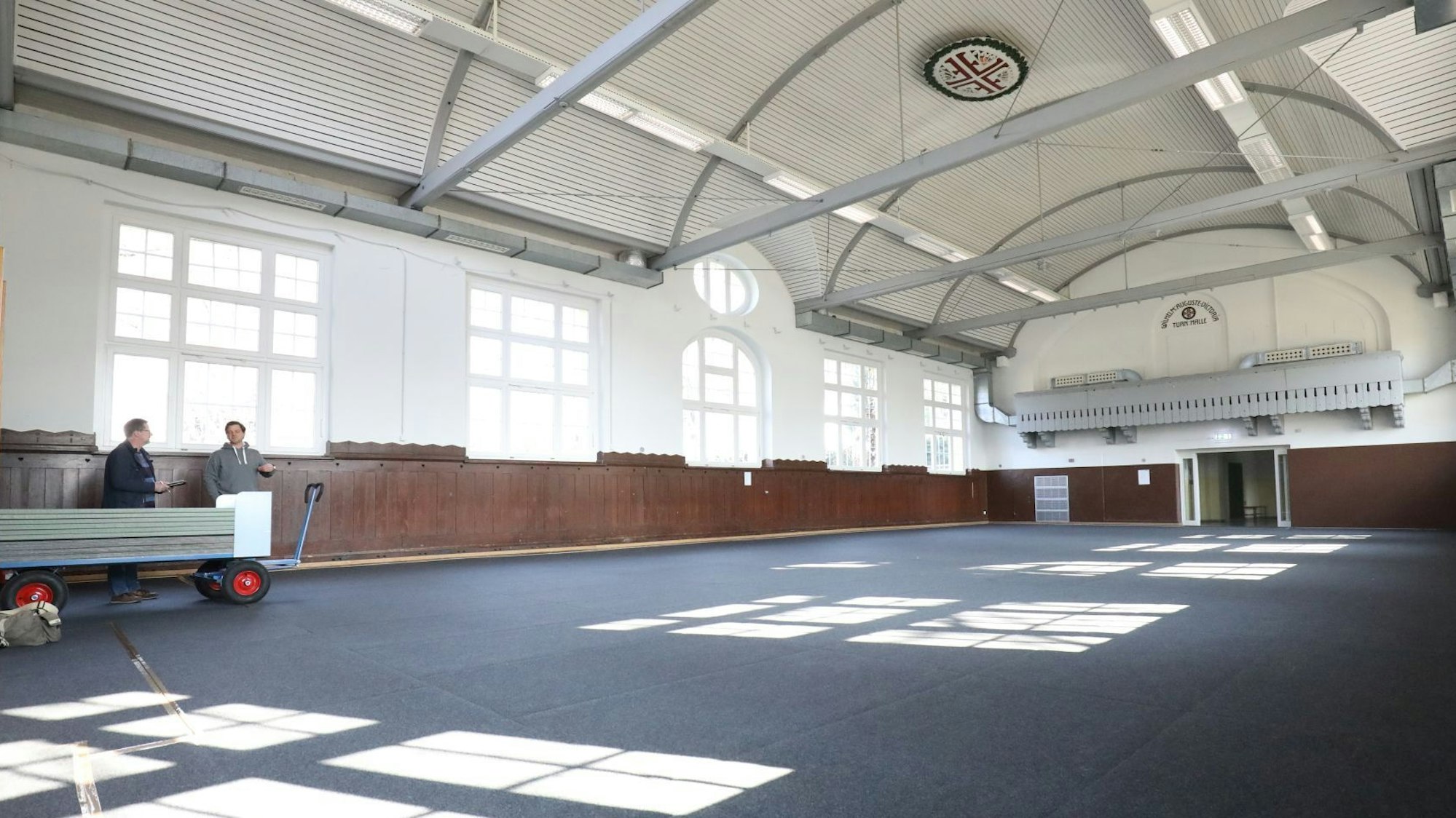 In einer fast leeren Turnhalle mit blauem Boden stehen am linken Rand zwei Personen.