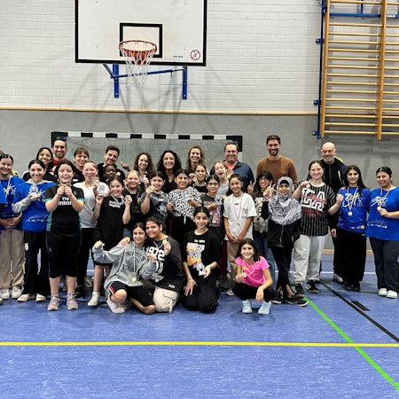 In einer Turnhalle posieren Schülerinnen mit Lehrer mit einer jungen Frau.