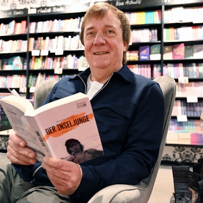 Ein Mann sitzt in einer Buchhandlung in einem Sessel. In der Hand hält er seinen eigenen Roman mit dem Titel „Der Inseljunge“.