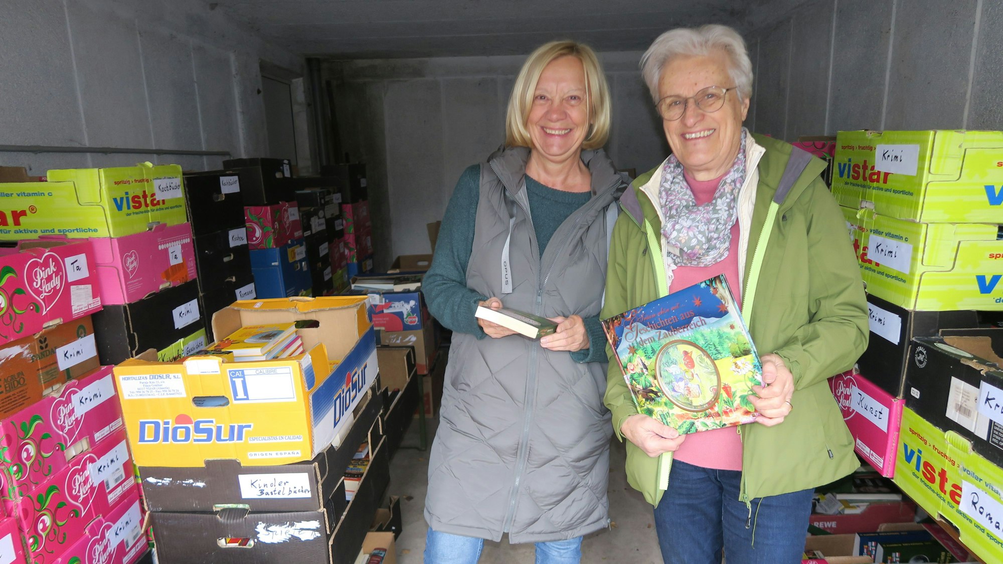 Zwei Frauen stehen in einer Garage inmitten von Kartons mit Büchern.