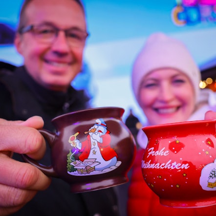 Ein Paar hält zwei Tassen mit heißem Glühwein kurz nach der Eröffnung eines Weihnachtsmarktes in den Händen und stößt an.