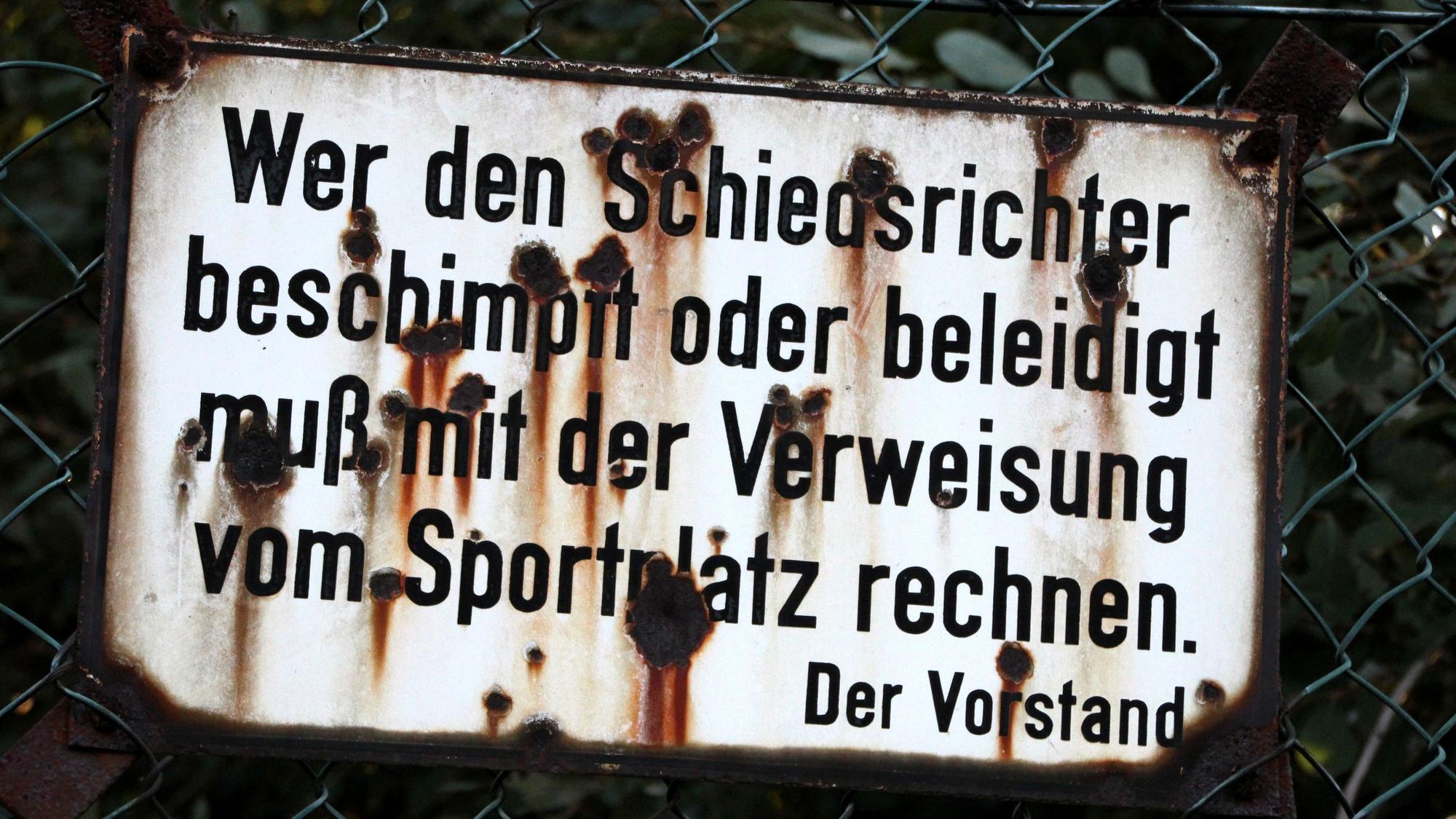 „Wer den Schiedsrichter beschimpft oder beleidigt, muß mit der Verweisung vom Sportplatz rechnen. Der Vorstand“ steht auf einem Schild an einem Sportplatz, das von Rost zerfressen ist.