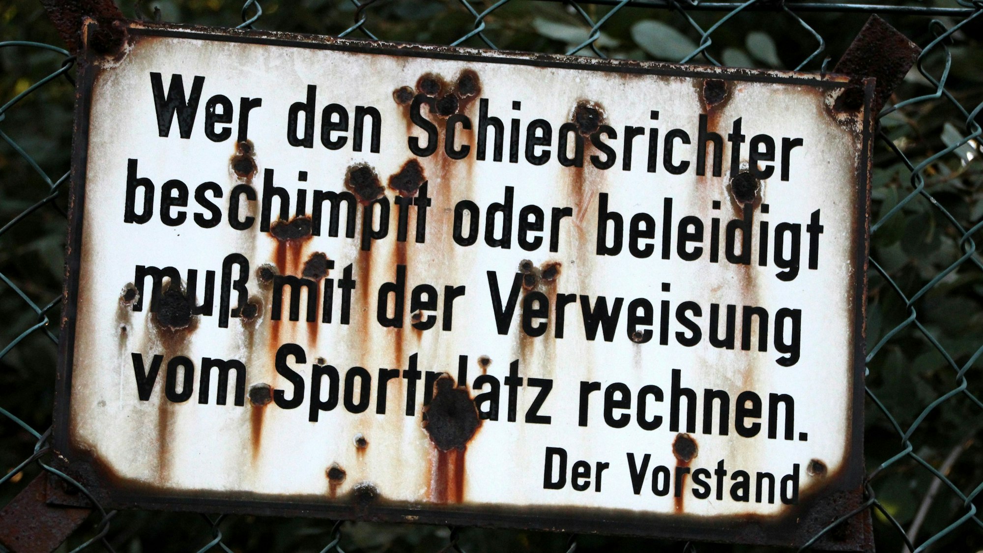„Wer den Schiedsrichter beschimpft oder beleidigt, muß mit der Verweisung vom Sportplatz rechnen. Der Vorstand“ steht auf einem Schild an einem Sportplatz, das von Rost zerfressen ist.