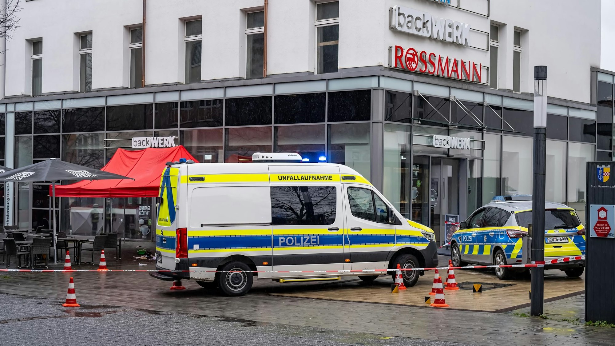 Zwei Polizeiautos stehen vor einem Backgeschäft. Auch Absperrband und ein Pavillon sind zu sehen.
