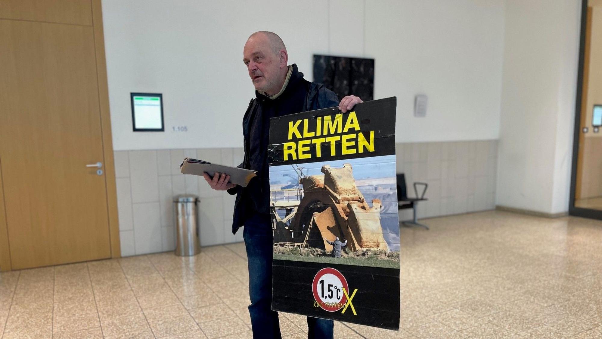 Klima-Aktivist Winfried Bernhard in Düsseldorf vor Gericht. Auf seinem Protestplakat: Das umstrittene Foto, das ihn vor einem Tagebaubagger zeigt. Das als Pressefoto des Jahres ausgezeichnete Bild zeigt ihn selbst, wie er vor einem Braunkohlebagger im Rheinischen Revier kniet.