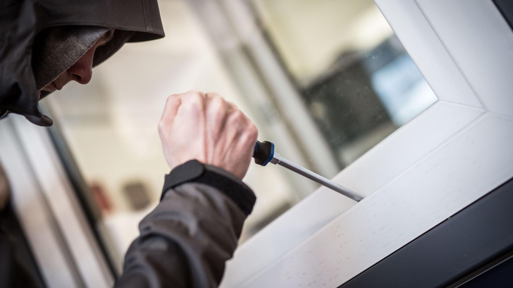 Das Bild zeigt eine vermummte Person, die mit einem Schraubenzieher versucht ein Fenster aufzuhebeln.