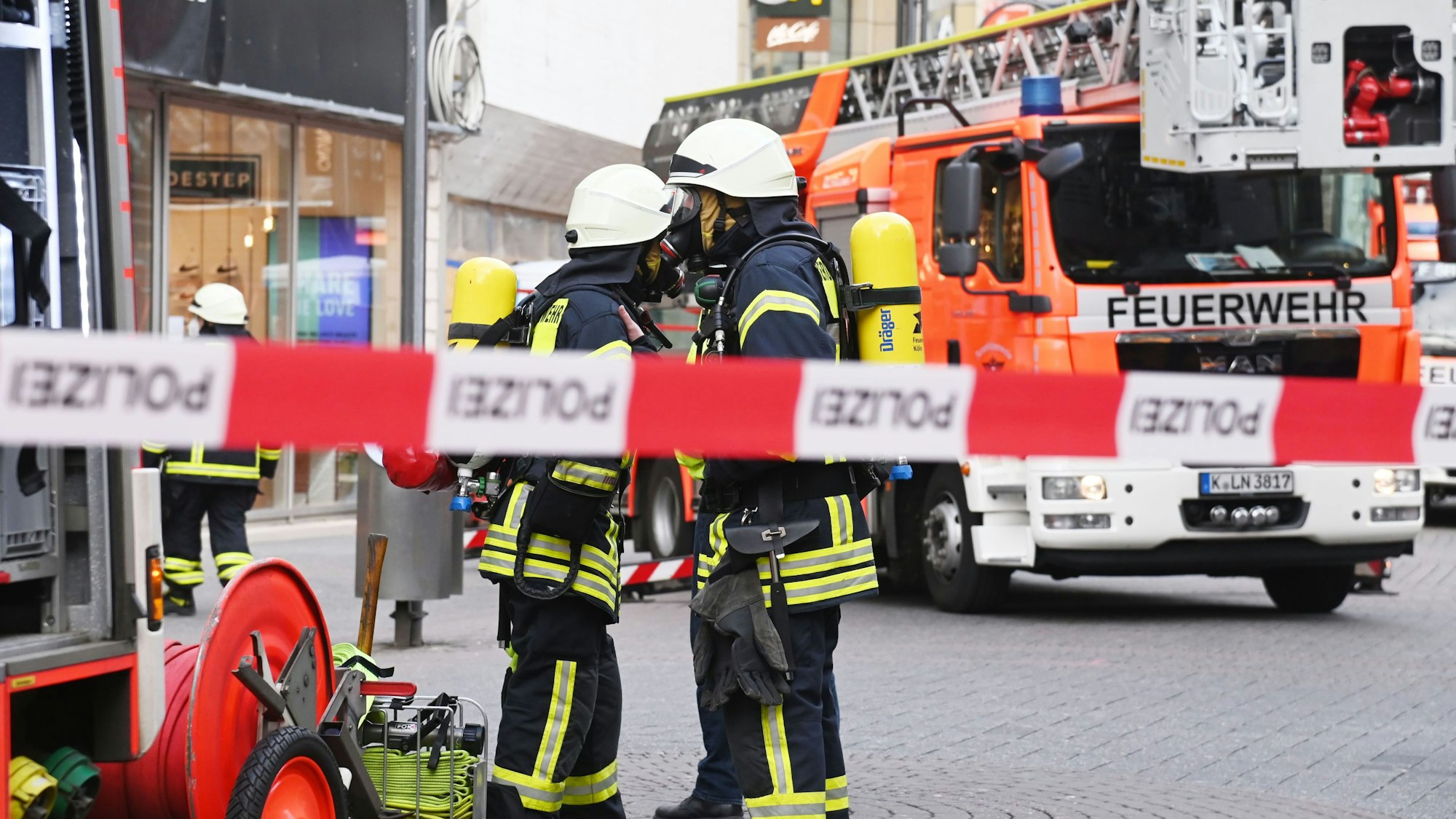Feuerwehrleute stehen im Anzug hinter einer Polizeiabsperrung, rechts im Bild steht ein Feuerwehrauto.