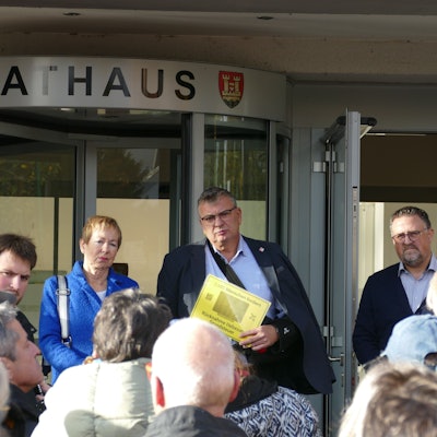 Vor dem Eingang zum Rathaus steht ein Mann, der eine gelbe Seite in der Hand hält. Vor ihm stehen mehrere Menschen, neben ihm eine ernste Frau mit kurzen Haaren.