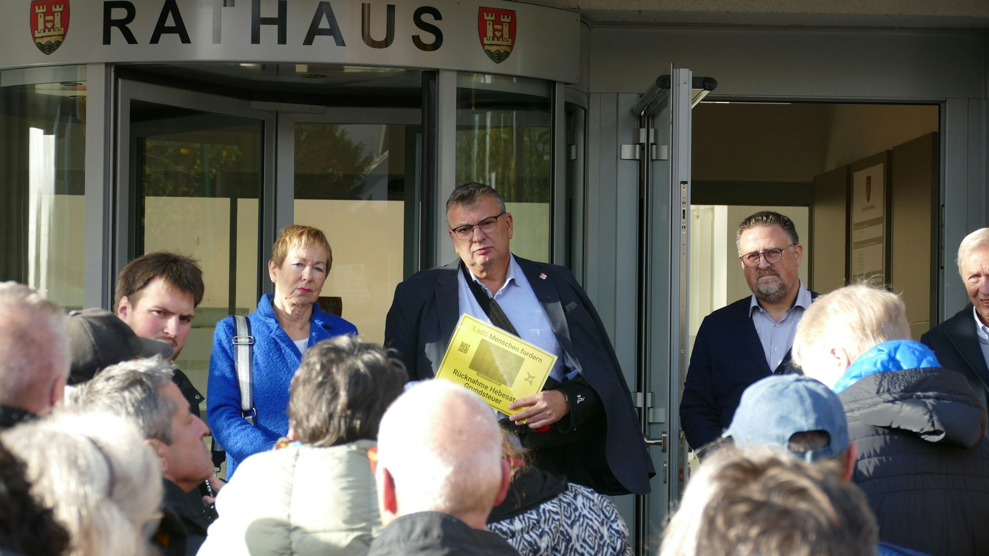 Vor dem Eingang zum Rathaus steht ein Mann, der eine gelbe Seite in der Hand hält. Vor ihm stehen mehrere Menschen, neben ihm eine ernste Frau mit kurzen Haaren.