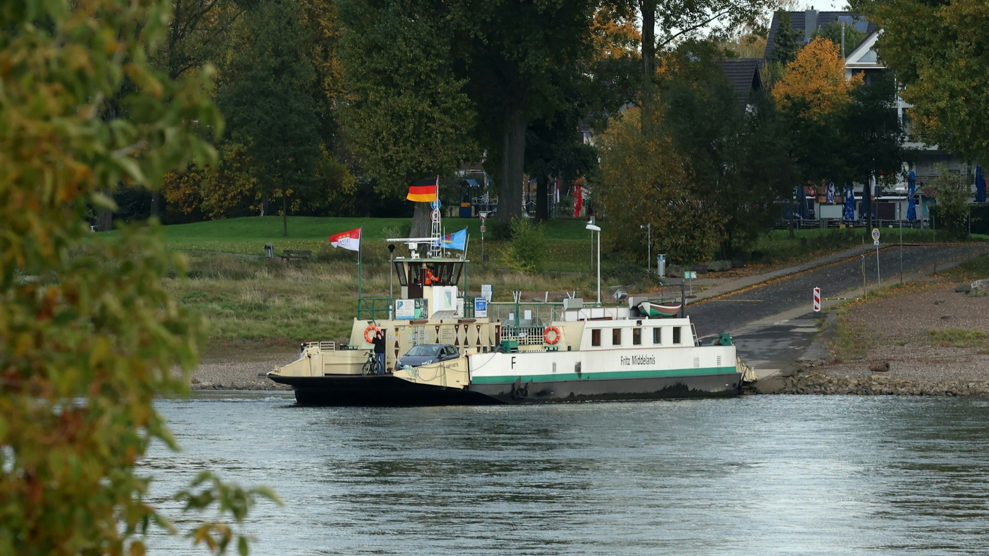 Die Rheinfähre „Fritz Middelanis“ liegt am Anlieger in Leverkusen-Hitdorf. Drumherum sind herbstlich gefärbte Bäume zu sehen.