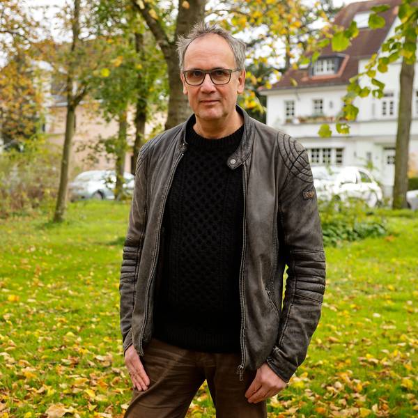 Schriftsteller Volker Kutscher steht im Klettenbergpark. Er trägt einen schwarzen Pullover und eine Lederjacke und blickt in die Kamera.