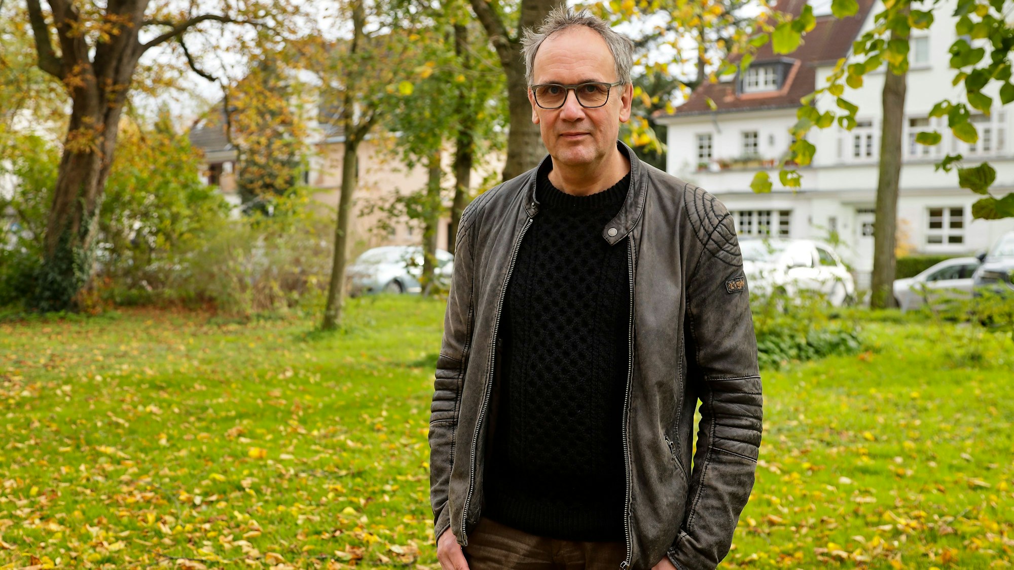 Schriftsteller Volker Kutscher steht im Klettenbergpark. Er trägt einen schwarzen Pullover und eine Lederjacke und blickt in die Kamera.