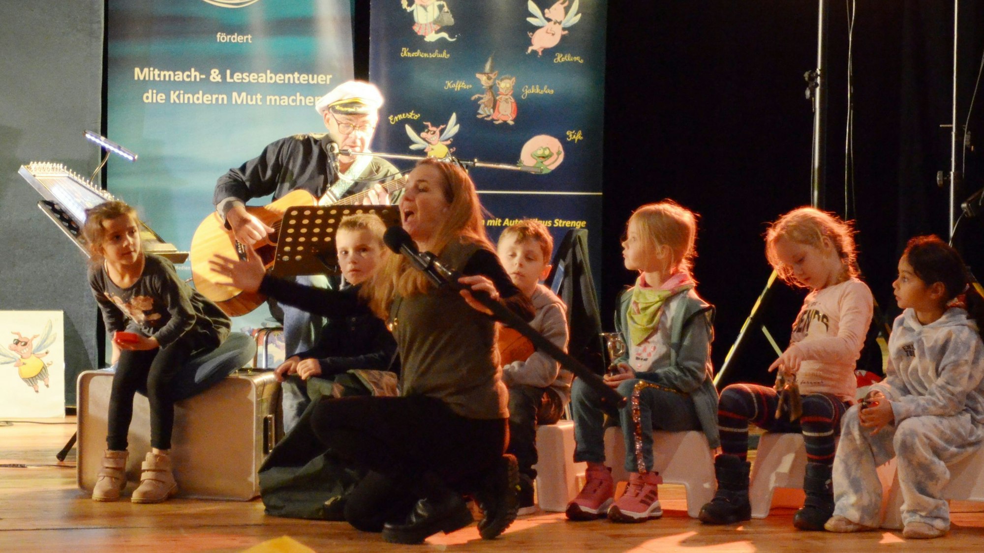 Die Moderatorin des Stücks „Der Kinderfresser kommt“, Andrea Herrmann, gibt auf der Euskirchener Theaterbühne Anweisungen. Hinter ihr sitzen Kinder und ein Gitarrist.